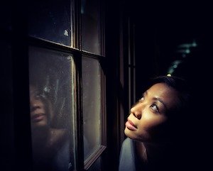 Black woman in dark room looks at her reflection in a window